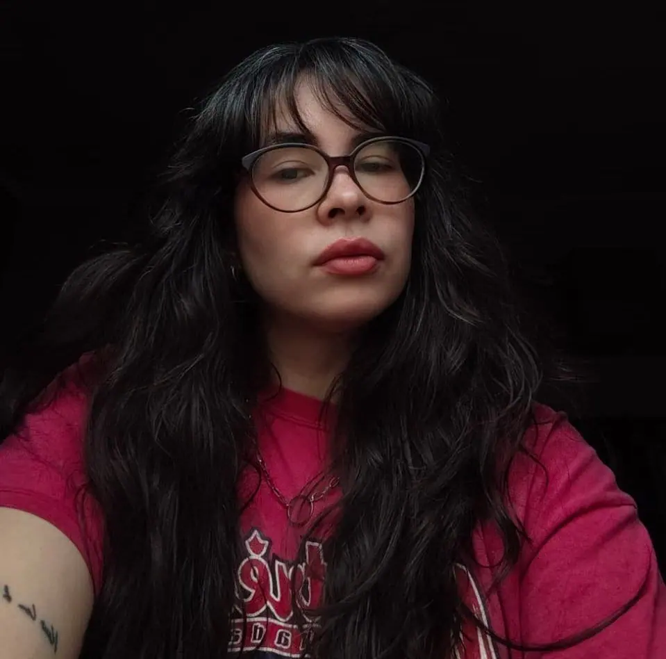 Manuela Justel, de óculos, cabelo longo e camiseta vermelha, sobre fundo escuro